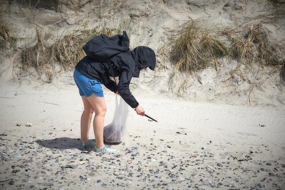 Person wearing light blue shorts, a rain jacket with a hood, and mint green barefeet shoes carrying a rubbish bag and a backpack picking up plastic in front of a sand dune with a pair of barbecue forceps.