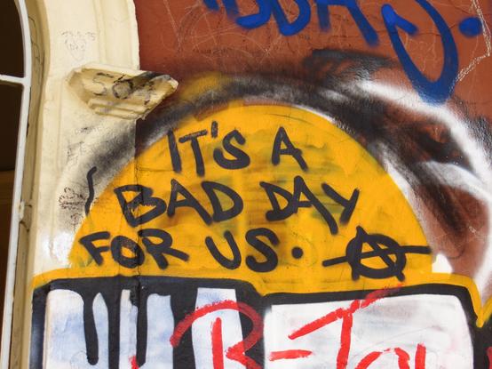 Eine mit Spraydosen besprühte Wand mit unterschiedlichen, abgeschnittenen Textteilen. Im Zentrum des Fotos steht in schwarzen Großbuchstaben auf einem hochkant gelben halbrund besprühtem Hintergrund: it's a bad day for us. daneben das Anarchie-Symbol  (ein A innerhalb eines Kreises)