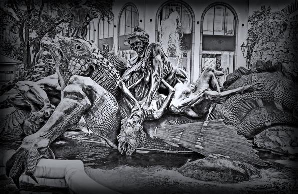 Stylized black and white photography of the "Fountain of Marriage" depicting various stages of a marriage in Nuremberg, Germany. In a large marble basin, a metal sculpture group is seen. Most of the basin is occupied by a giant lizard extending horizontally, with its head to the left and looking up into the skies. Its skin has countless dents. On the back of the lizard, a couple depicted as half-decomposed corpses is fighting. The man is lying on his back, his head hanging down and his hair almost touching the water surface. What remained of his eyes is looking fearful. He is trying (rather unsuccessfully) to stop the woman that is raised above him from strangling him.
