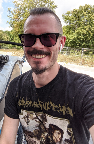 A man with an (what I like to think of as) Oliver-Queen/Green-Arrow-style beard and sunglasses grinning into the camera, airpods in and Mohawk hair bound back. He's sitting next to a jogger, apparently done with his half hour of running. The pic is cropped so the kid can't be seen.