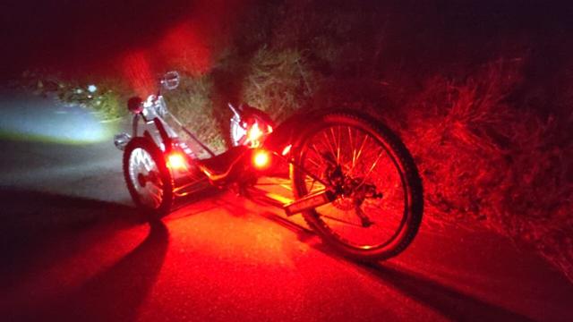 A rather low KMX recumbent trike askew from the back with four bright LED lights, two on the handle bar and two in the back of the backrest.