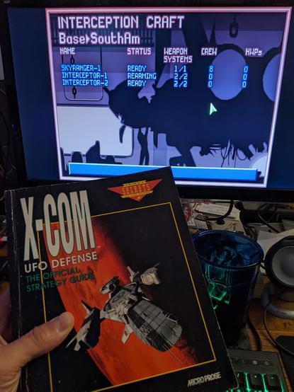 Paradroyd is holding an X-Com UFO Defense official strategy guide up to the camera.  In the background is a monitor playing the game and is on an "interception craft" screen for the SouthAM base.