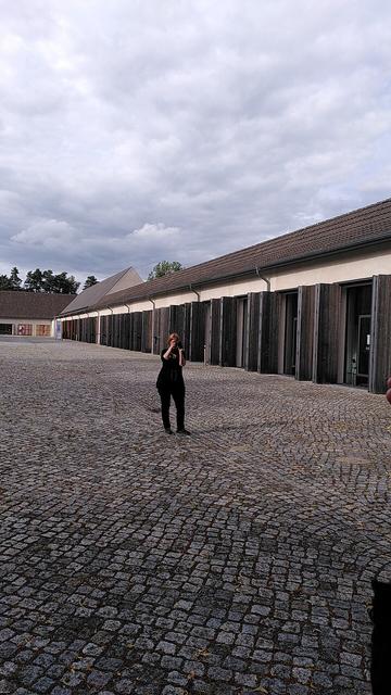 Rechts ehemalige Garagen, die heute Fenster haben und als Büros dienen. Auf einem gepflasterten Weg steht eine Frau in schwarzer Kleidung, die fotografiert.