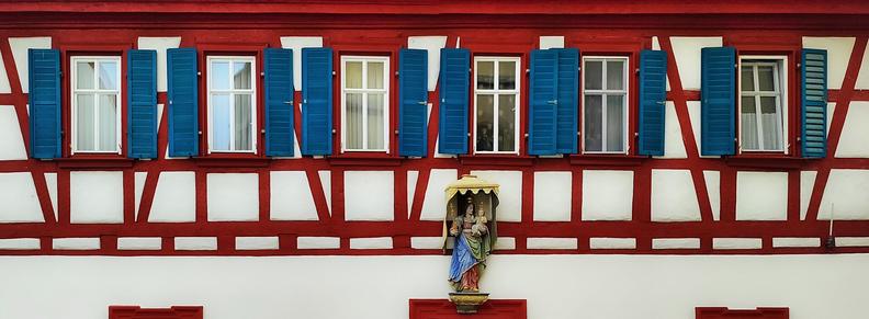 Upper half of the wall of a half-timbered house with white plaster. The timbers are crimson. At the top, from left to right, six subdivided windows with dark blue, wooden window shutters. The ground level of the wall isn't visible, apart from the lintels of the door and two windows, left and right of the door, respectively. Directly over the entrance, under a golden canopy, a statue of Mary holding Jesus. In her right hand, she is holding a sceptre. Jesus is holding an orb in his left hand. Both are wearing golden crowns.