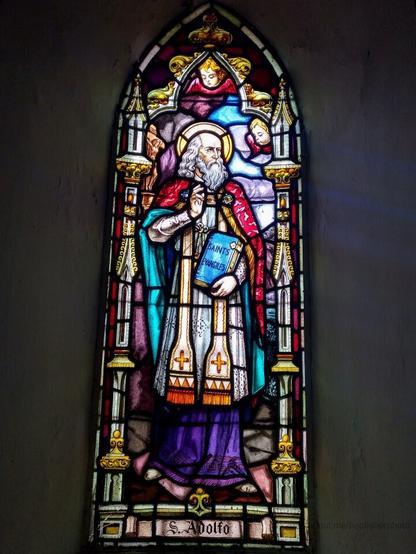 Photograph of a stained glass window, depicting a holy figure dressed in robes and holding a book of the bible, labelled "S Adolfo". There are cherubs floating around the figure.