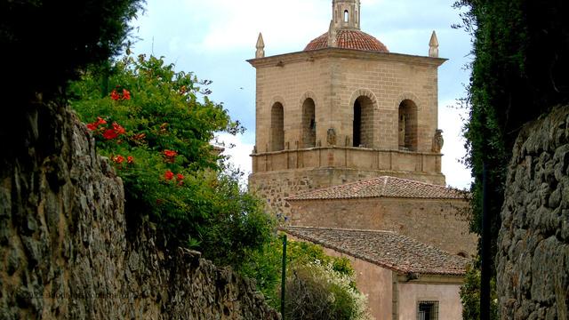 Photo looking down an alleyway in an old Spanish city, with tall old walls topped with lush green plants and bright red flowers. At the end of the alleyway is a light brown brick tower with arches and smaller decorative towers on top of it.