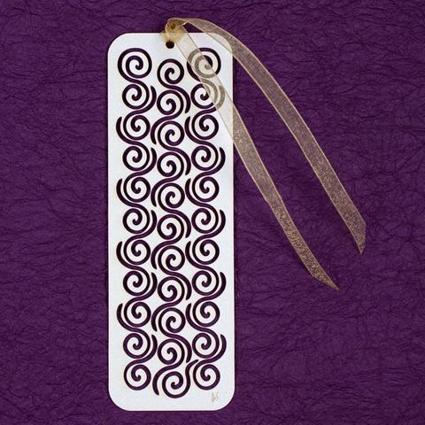 Bookmark with a pattern of repeating spirals with long, curvy tails cut from white stock with a metallic sheen, gauzy gold ribbon tied to one end, crinkly purple paper background.
