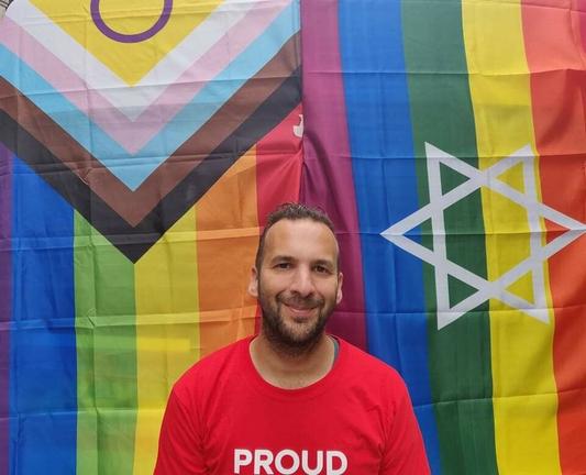 Zack in front of the intersectional flag and a rainbow star of David. He's wearing a red t-shirt with the word "Proud."