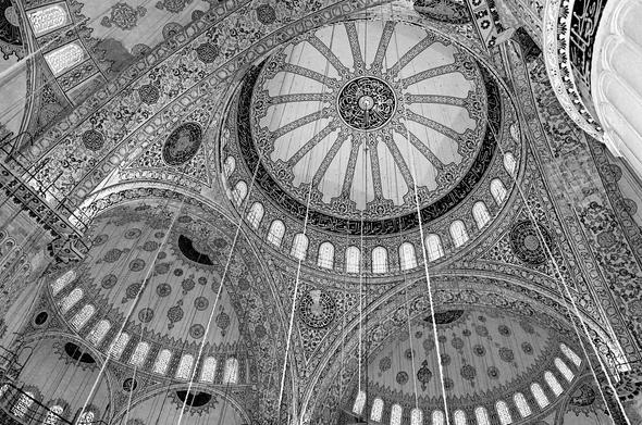 Black and white photo of an inside view up the central dome of the Sultan Ahmed Mosque (Blue Mosque) at Istanbul, Turkey. The whole interior is decorated with tiles that have floral ornaments and inscriptions, with dominant colors of blue and red. The upper parts of the mosque are lit by numerous stained glass windows. Running down from many points of the ceilings are suspension wires (meant for holding unadorned iron chandelier rings that are not in the view field of this photograph).