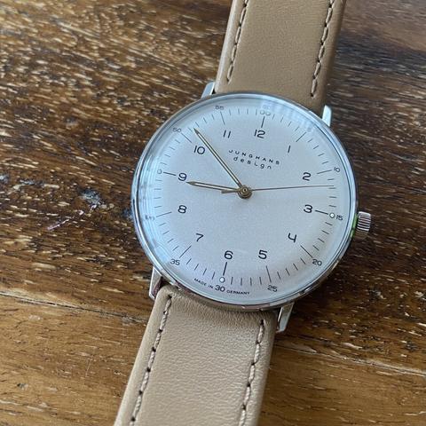 Eine Armbanduhr mit weißem Zifferblatt und beige farbenem Lederband liegt auf einem Holztisch.
