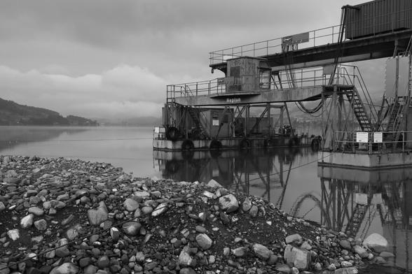 Schwarz-weiß Aufnahme eines Schwimmbaggers in Ufernähe. Spiegelung im Wasser. Im Vordergrund Kies/Schotter am Ufer im Hintergrund Wolken.