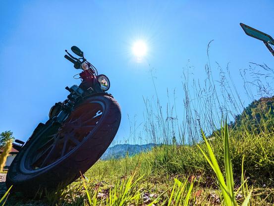 Motorrad vom Boden aus fotografiert, man sieht den Reifen und etwas grünes Gras. Im Hintergrund strahlend blauer Himmel und Sonnenschein sowie ein paar Berge.