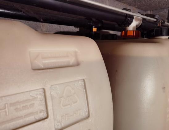 Two heating oil tanks in a basement  side by side. The oil level is visible through the translucent plastic tank walls. The level is right on the maximum fill line on the near tank. Above the tanks some of the piping is visible.
