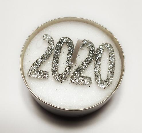 On a white background, a tealight in its aluminum cup, decorated with a glittery silver "2020". The wick is unlit.