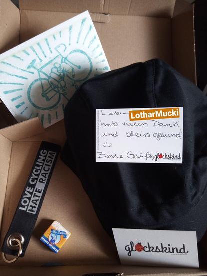 Ein åpend lütt Paket wo binnen liggt: ne Dankseggungskord för de Bestöllung, n Schlœdelanhanger worup steiht: "Love Cycling Hate Racism", n lütt Packen Bruuspulver, ne Visten- un ne Postkord un ne handneiht Radfohrklott ut Boomwull.