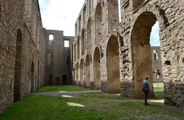 The inside of a three-story ruin. The walls still seem pretty intact, but the floor are gone and on the ground grass is growing. In the walls are very large arched windows and other openings. To the right a person is standing looking through one of the arches.