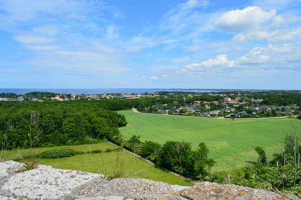 A view from atop of the castle walls, looking towards the city of Borgholm in the distance. In the front are lots of trees and grass and the sky is very blue. In the distance the ocean is visible.