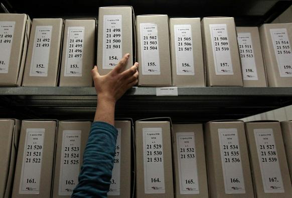 "Une main est tendue, depuis le bas de la photo, vers une rangée de boîtes d'archives, avec des étiquettes comportant des côtes, et s'apprête à en saisir une. "