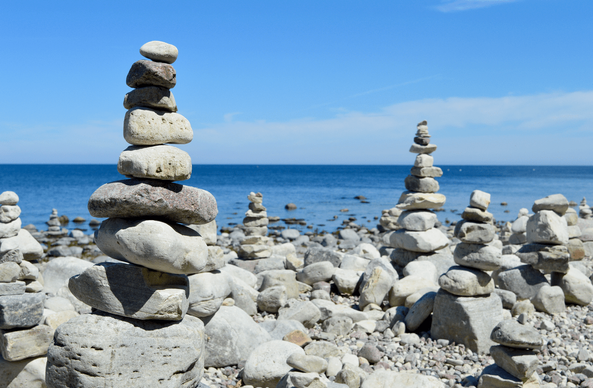 A shingle beach with many stacks of single rocks stacked on top of each other. In between these stacks of rocks are more rocks and small pebbles. The photo is taken close to one stack on the left. To the right, a little further away, are more stacks which are slightly out of focus. In the distance the ocean is visible.