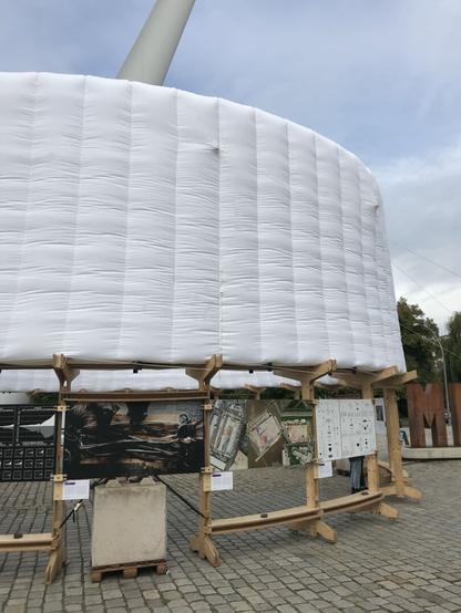 An outside exhibition, a circular wooden construction holding images and above a large white tube that extends the exhibition construction above