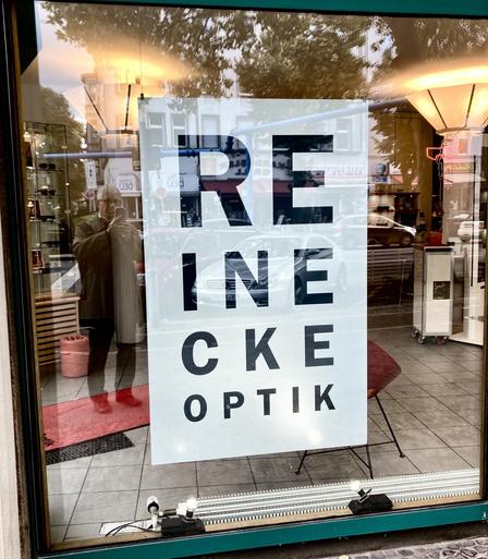 Firmenschild, bei dem der Text „Reineckeoptik“ auf mehrere Zeilen untereinander mit immer kleineren Buchstaben verteilt ist - wie die Sehtesttafeln beim Optiker.
