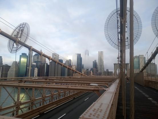 A photograph of the Manhattan skyline early in the morning of September 12, 2022, from the middle of the Brooklyn Bridge. The fog is beginning to set in and the Freedom Tower is almost entirely obscured, but the tall skyscrapers in front of it are clear.