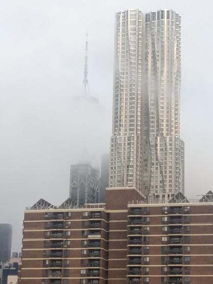 A photo of the Freedom Tower from close to the Manhattan exit of the Brooklyn Bridge. The morning fog is heavy, and despite being close to the Freedom Tower for the photo, the tallest building in NYC is almost entirely obscured save for its spire.