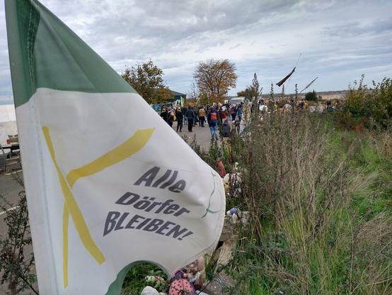 Alle Dörfer bleiben Fahne auf Erdwall. Im Hintergrund Menschen beim Auftakt Dorfspaziergang Lützerath