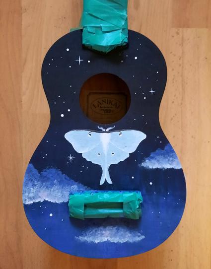 Der Körper einer Ukulele, Hals und Sattel sind mit Malerkrepp abgeklebt. Ein Farbverlauf von Schwarz zu Blau zieht sich von oben nach unten über den Körper. Auf dem unteren Teil sind drei kleine Wolken aufgemalt, über ihnen, auf dem schwarzen Teil, funkeln Sterne. Direkt unter dem Schallloch schwebt eine Luna Motte. Ihre Flügel sind blassgrün.