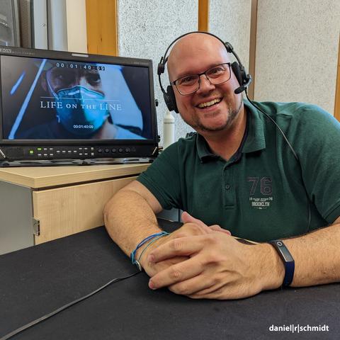 Der Studiosprecher Daniel R. Schmidt kniet vor einem Bildschirm. Er hat ein professionelles Studioheadset auf. Auf dem Bildschirm ist ein Arzt in einer blauen FFP2-Maske zu sehen. Das Titelbild zeigt zudem den Schriftzug "Life on the Line".