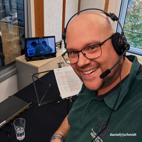 Der Studiosprecher Daniel R. Schmidt steht vor einem Bildschirm. Er hat ein professionelles Studioheadset auf. Auf dem Bildschirm ist eine Szene aus der TV-Serie "Life on the line" zu sehen.