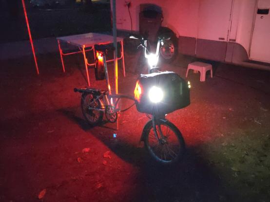 Brompton Faltrad von schräg vorne bei Nacht mit Knog Plus Akku-Leuchten an allen Ecken der Fronttasche (vorne weiß, hinten rot), eine Knog Copper Akku-Leuchte am Lenkkopf und eine Knog Blinder Akku-Leuchte an der Sattelstange — zusätzlich zu den Dynamo-Lichtern des Bromptons, die auf dem Bild aber nur noch im Standlicht-Modus leuchten.