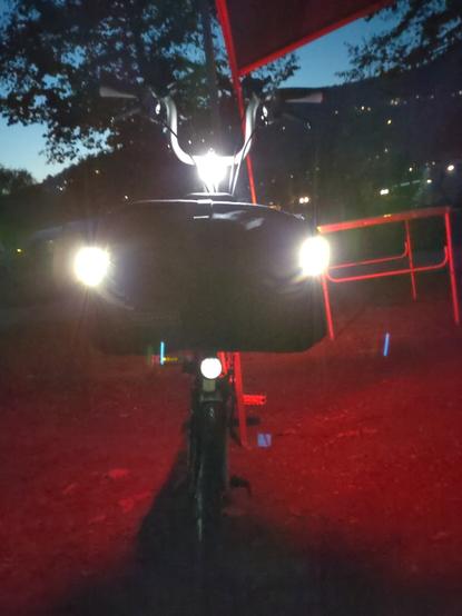 Brompton Faltrad von vorne bei Nacht mit Knog Plus Akku-Leuchten an den Ecken der Fronttasche (man sieht nur die beiden nach vorne weiß leuchtenden), eine Knog Copper Akku-Leuchte am Lenkkopf — zusätzlich zum Dynamo-Scheinwerfer des Bromptons, ein Busch und Müller IQ-X, welcher auf dem Bild aber nur noch im Standlicht-Modus leuchtet. Die vier Frontleuchten bilden ein auf der unteren Ecke stehendes nahezu symmetrisches Trapez.