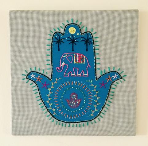 An embroidered piece showing a blue hamsa with an elephant , palm trees and moon, a mandala and floral stitches