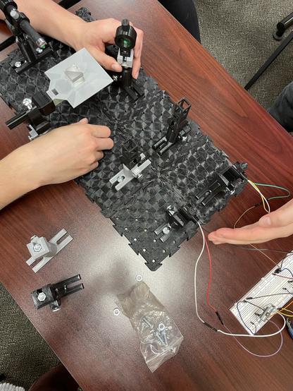 A photo from above. On a dark-wood table a black 3D printed optical breadboard holds mounts for a laser pointer, beam splitter cube, mirror, two cuvette holders, and two photodiode detectors. The optical breadboard is oriented diagonally in the photo. In the top left a student’s hands manipulate the beam splitter cube and mirror to direct green laser light toward a detector. In the bottom right beyond the detectors wires connect to an electronics breadboard with a circuit for each detector. Another student’s hand is cupped behind the detector to monitor the location of the laser beam. Scattered around in the table are extra mounts and a small clear plastic baggie of nuts and bolts.