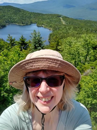 Head shot of Erin Brenner in a hat and sunglasses with a view of a lake and mountains behind her.