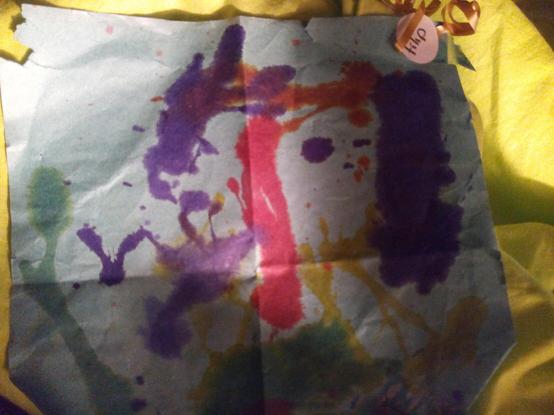 A photo of a kid's painting; various colours on the paper; quite possibly made by spreading liquid water colours by blowing them around with a straw?
I can see a green tree, a purple bird, a green and yellow bush, a pink slide, a purple puddle or lake.