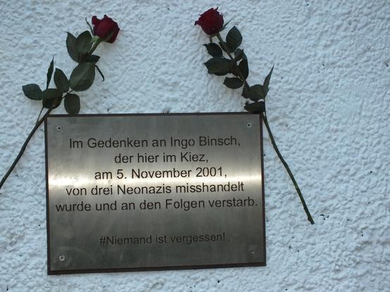 Metallgedenktafel an einer weißen Wand:
Inschrift:
Im Gedenekan an Ingo Binsch, der hier im Kiez, am 5. November 2001, von drei Neonazis misshandelt wurde un an den Folgen verstarb.

#niemand ist vergessen

Dazu zwei Rosen