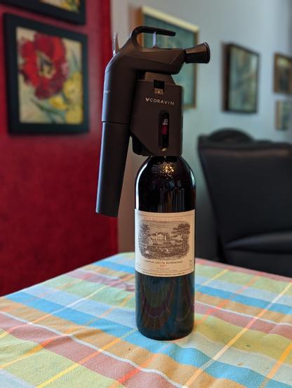A bottle of 1997 Chateau Lafite Rothschild with a Coravin wine preserving system attached to the neck sits on a dining table, against a slightly blurred background.