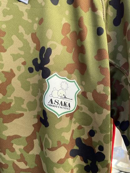 Asaka Shooting Venue: camouflage pattern t-shirt with a cartoon dandelion character lining up a shot with an olympic rifle