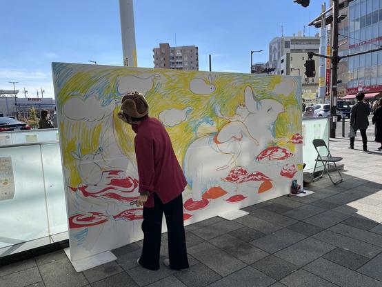 An artist draws a whimsical picture of rabbit like creatures on a large board outside a train station