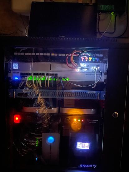 A picture of my basement networking rack, in the dark, with lots of status and indicator lights visible.