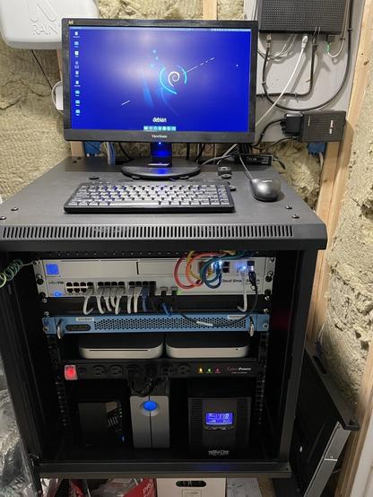 A pic of my black metal networking rack in a basement room with insulation on the walls and bare joists visible. The rack is filled from top to bottom with network hardware, servers, and disk drive enclosures. On the top is a LCD monitor, keyboard, and mouse at approximately standing desk height.