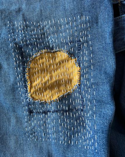 The blue denim leg of a pair of overalls, with a yellow patch, and fine white sashiko-like (not traditional) stitches covering the area.