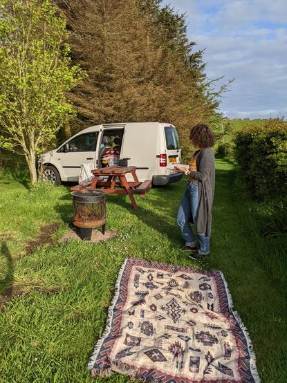 Van set up in a camping area with picnic blanket, fire pit and picnic table. Incredibly beautiful woman standing looking away from camera