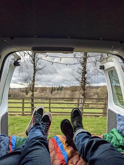 Two people's crossed legs in view and feet hanging out of open back Doors of a van. Looking out into a field with forests in the distance.