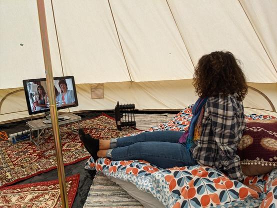 Exceptionally beautiful woman sitting on an inflatable bed watching TV inside a huge tent. Floor is covered in rugs. Electric heater in view.