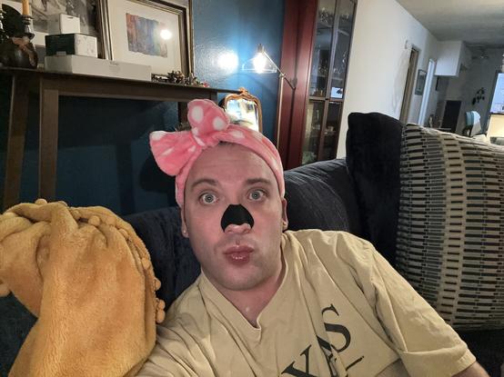 Man with black charcoal nose strip on whose hair is pushed back by a plushy, pink polka-dotted head scrunchy making a dramatic duck face.