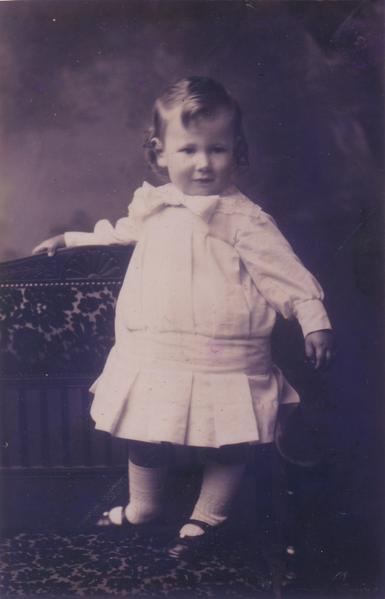 Photo of a young Harry Laurie as a toddler, in a white cotton dress, which was common for all genders 100 years ago. He has curly hair and white socks with black leather shoes and is standing at the back of a sofa with a pattern on it.