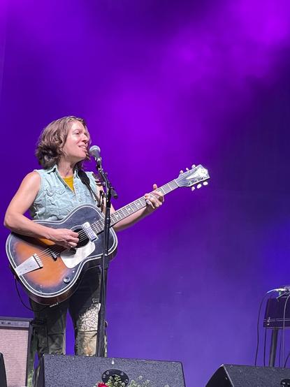 A woman stands on stage, playing a guitar.